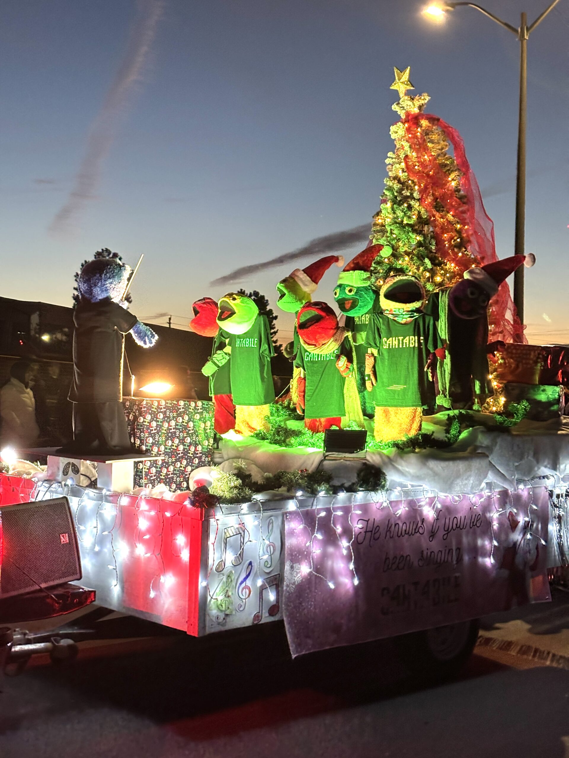 Christmas parade float featuring colorful puppet characters dressed as choir singers, a conductor, and a large decorated Christmas tree.