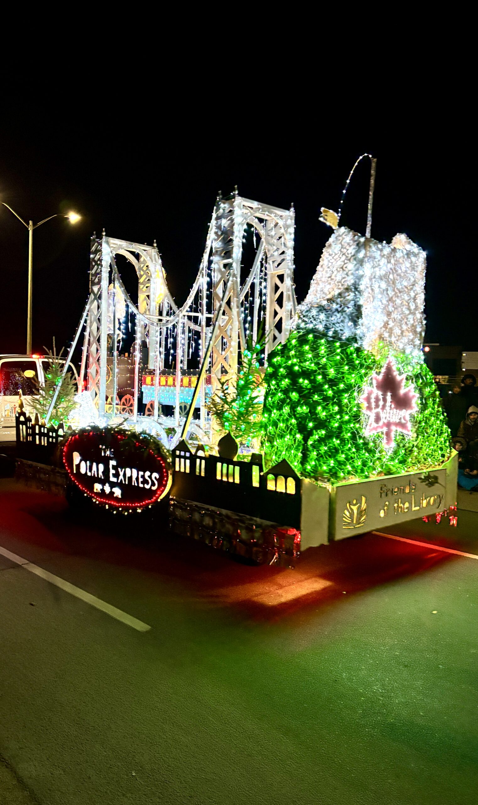Polar Express-themed Christmas parade float featuring a glowing bridge, lit-up train, snowy mountain, and colorful holiday lights.