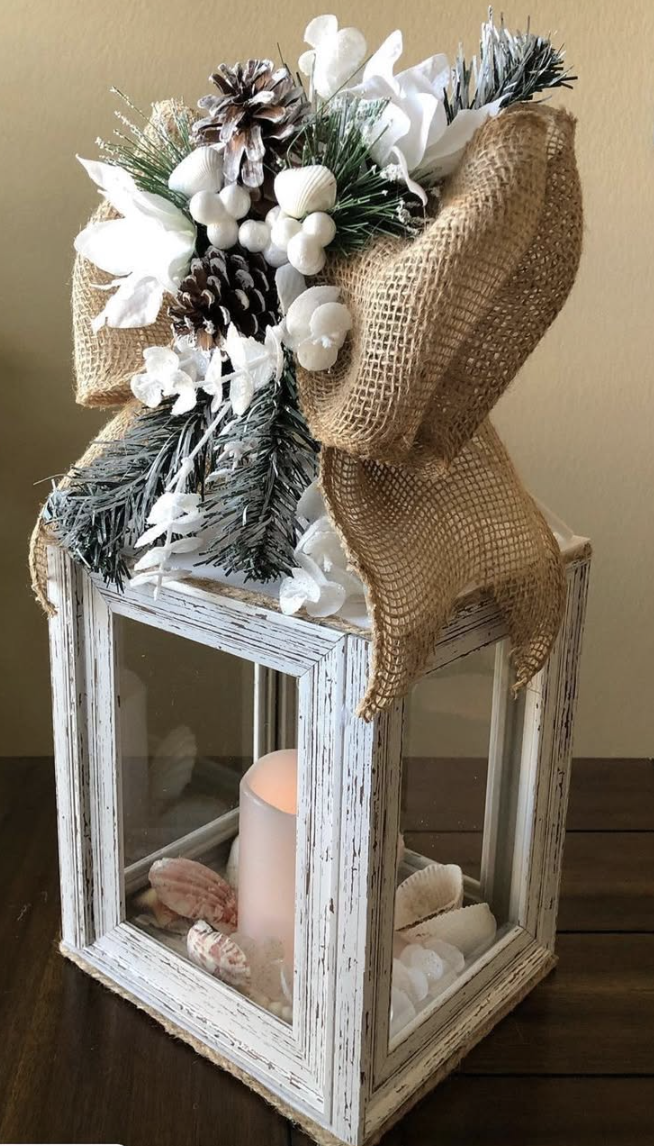 Rustic white lantern decorated with burlap bow, pinecones, frosted greenery, and a glowing candle for Christmas décor.