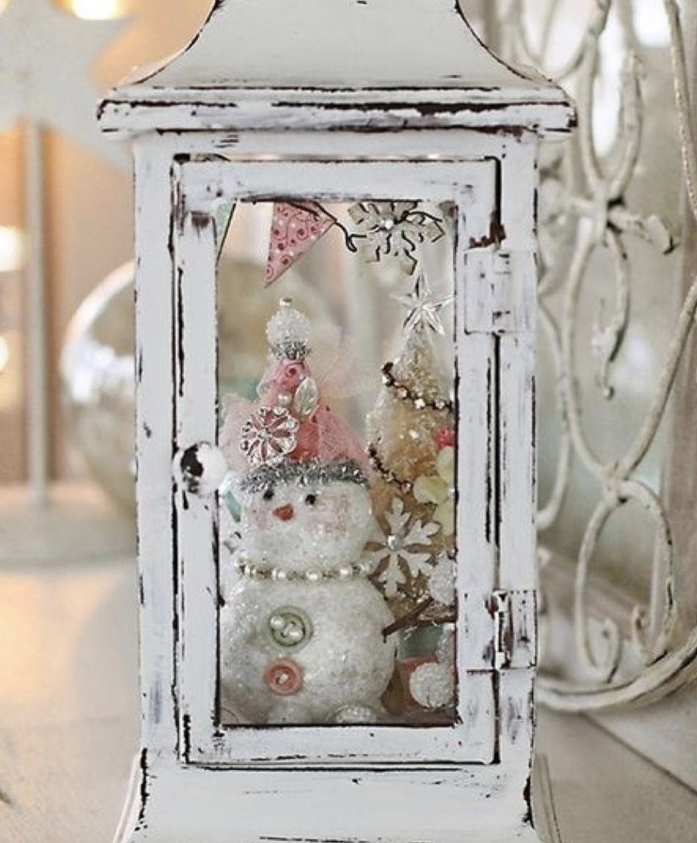 Distressed white shabby chic lantern filled with a glitter snowman figurine, mini Christmas trees, and ornaments for holiday décor.
