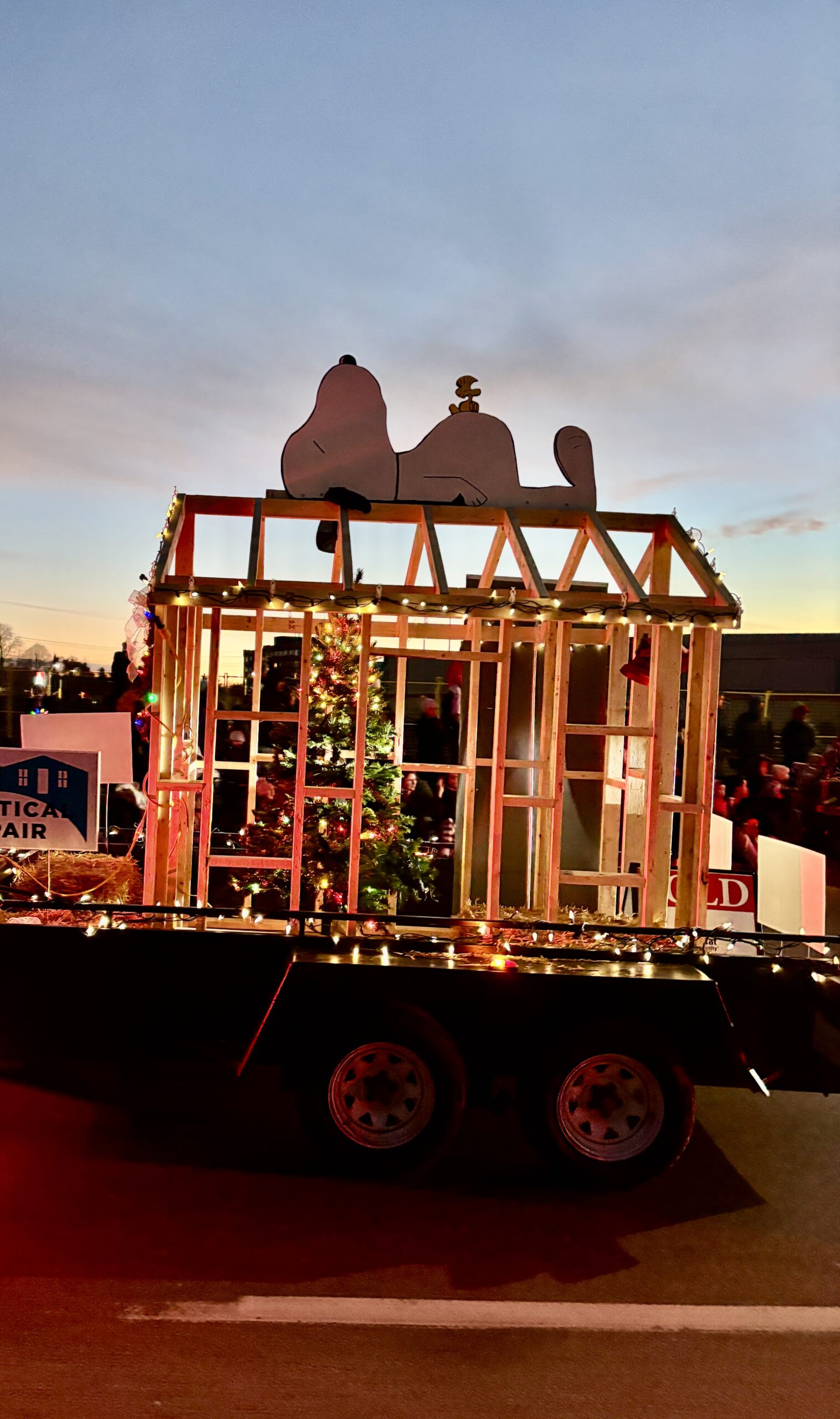 Christmas parade float featuring a framed wooden house structure decorated with lights, a Christmas tree, and a large Snoopy and Woodstock cutout on top.