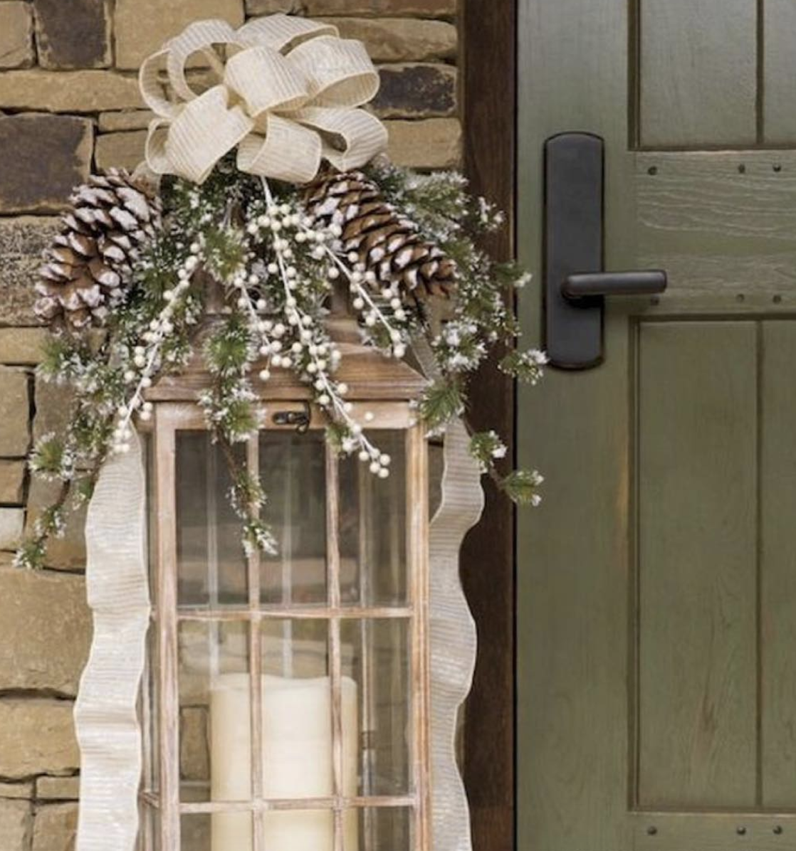 Tall rustic outdoor lantern with candles, topped with pinecones, frosted greenery, and a large ivory bow for Christmas décor.