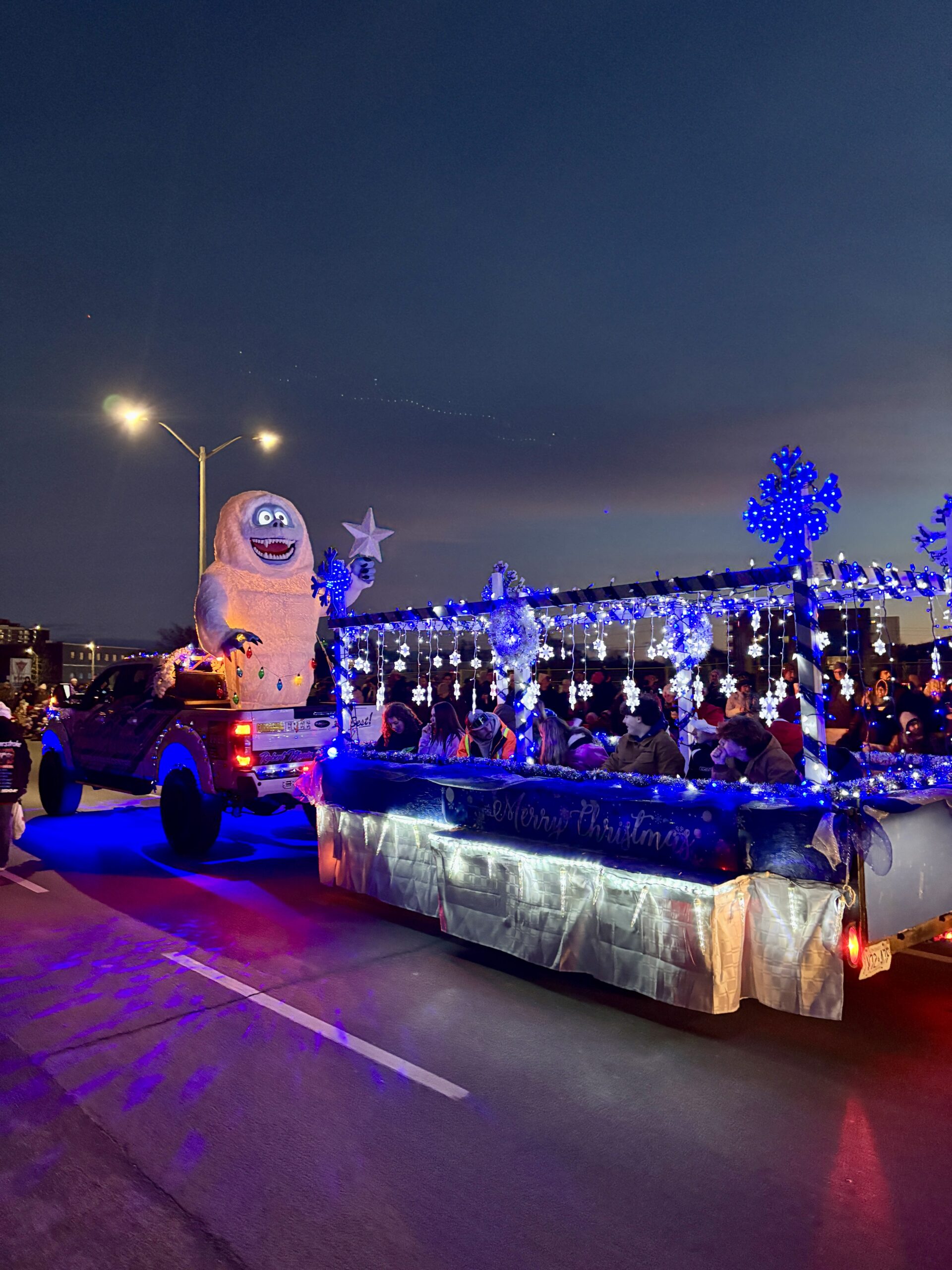 Christmas parade float with a large smiling yeti figure pulling a winter wonderland trailer decorated with blue lights, snowflakes, and holiday characters.