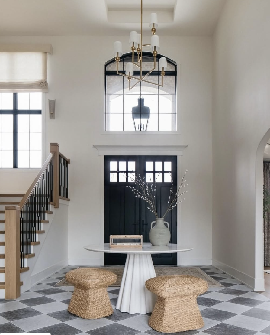 Modern brass chandelier hanging in a two-story foyer with black front doors, checkered tile floor, and minimalist entry table and stools
