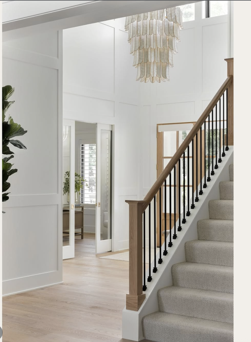 Soft neutral chandelier hanging in a two-story foyer with light wood floors, black iron stair railing, and white paneled walls