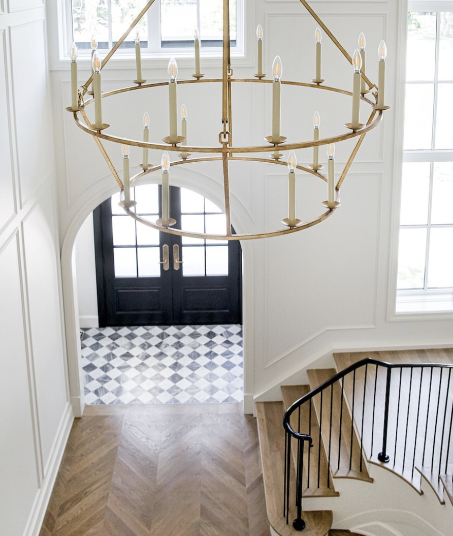 Oversized candle ring chandelier hanging in a two-story foyer with black front doors, herringbone wood floors, and curved staircase railing