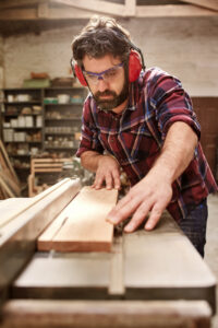 Bearded carpenter working on his studio, cutting a wooden plank with a circular saw, and wearing sfatey goggles and earmuffs - Safety Equipment You need when Doing DIY in the Home