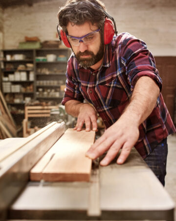 Bearded carpenter working on his studio, cutting a wooden plank with a circular saw, and wearing sfatey goggles and earmuffs - Safety Equipment You need when Doing DIY in the Home
