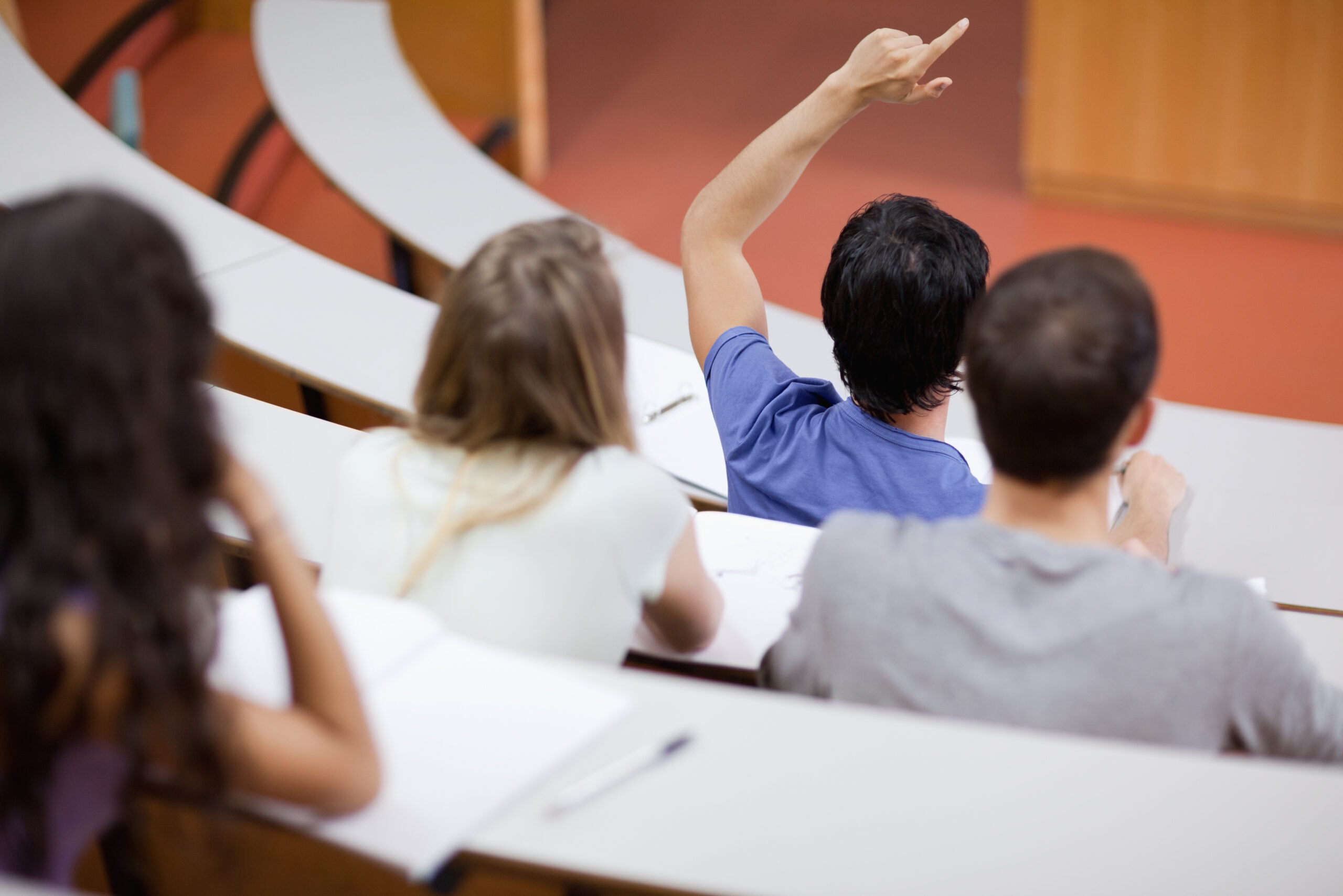 Young student raising his hand while his classmates are taking notes in an amphithater