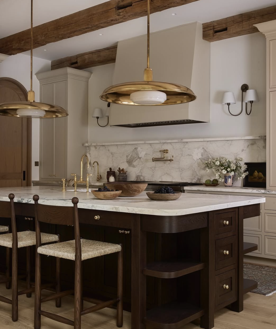 Kitchen with a statement plaster range hood and marble slab backsplash, warm wood island, brass lighting, and exposed wood ceiling beams.