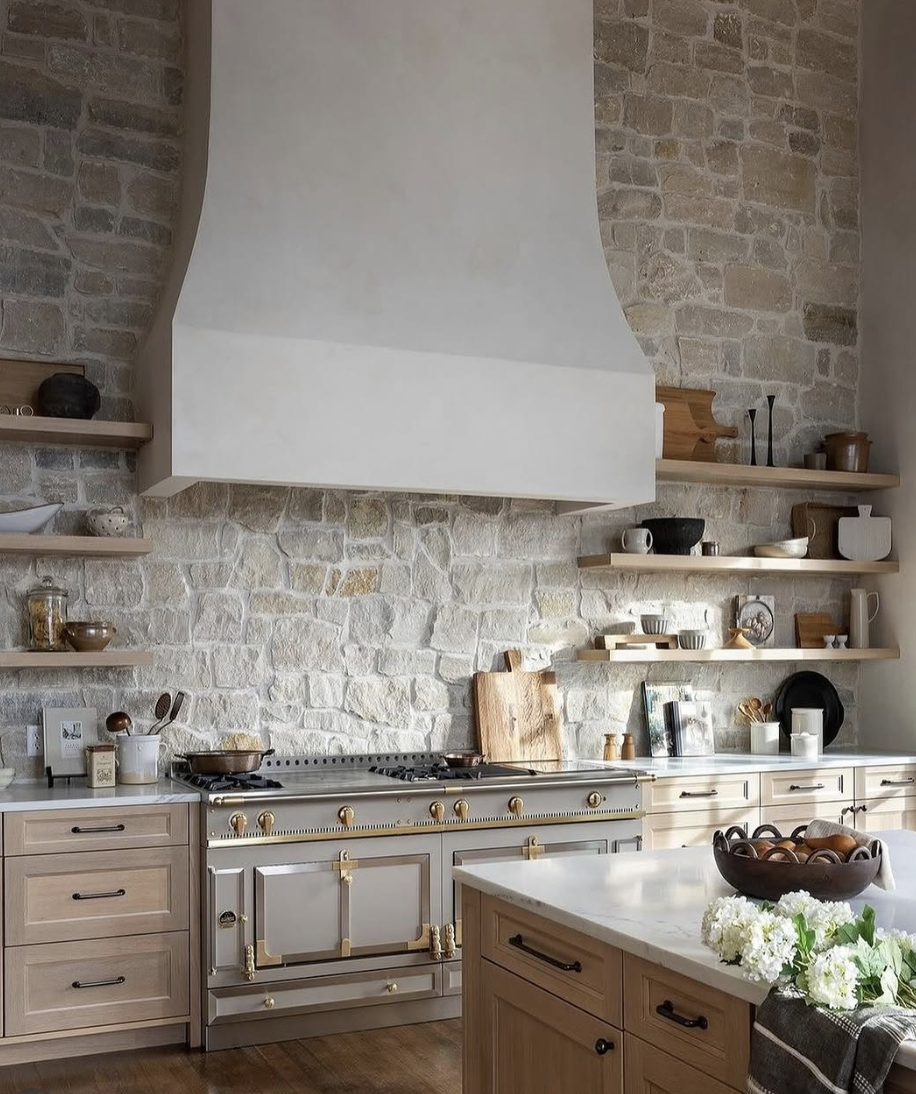 Farmhouse kitchen with a full-height natural stone slab backsplash, custom plaster range hood, open wood shelving, and a statement gas range.