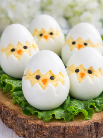 Easter deviled egg chicks arranged on lettuce over a rustic wooden board, decorated with carrot beaks and peppercorn eyes.