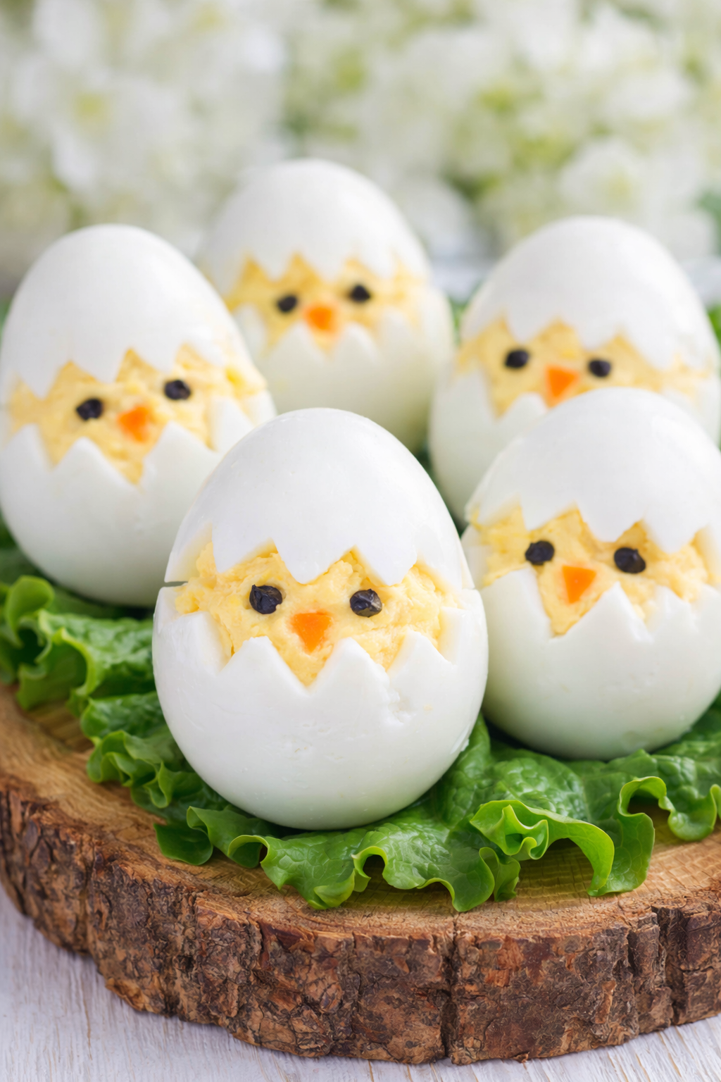 Easter deviled egg chicks arranged on lettuce over a rustic wooden board, decorated with carrot beaks and peppercorn eyes.