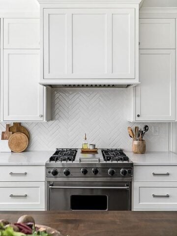 White shaker cabinets, beautiful marble countertop. White wood paneled range hood, white herringbone backsplash tiles above a stainless steel oven range.