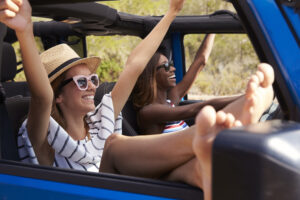 Two Female Friends Driving Open Top Car On Country Road -- Spring is the perfect time to get out and explore new destinations. Here are 4 reasons why you should take a road trip this spring