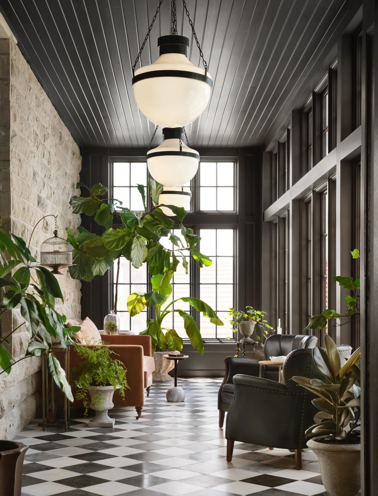 Sunroom living space by Joanna Gaines featuring checkerboard floors, oversized pendant lighting, leather chairs, stone walls, and lush greenery.