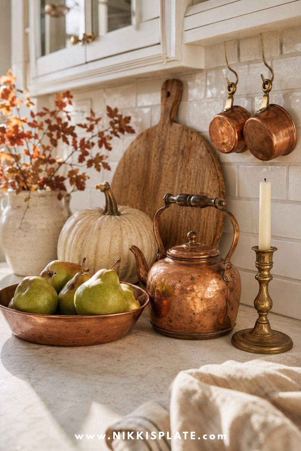 Vintage copper kettle, copper bowl of pears and aged brass candlestick styled with warm wood, a cream pumpkin and autumn foliage in a light farmhouse kitchen.