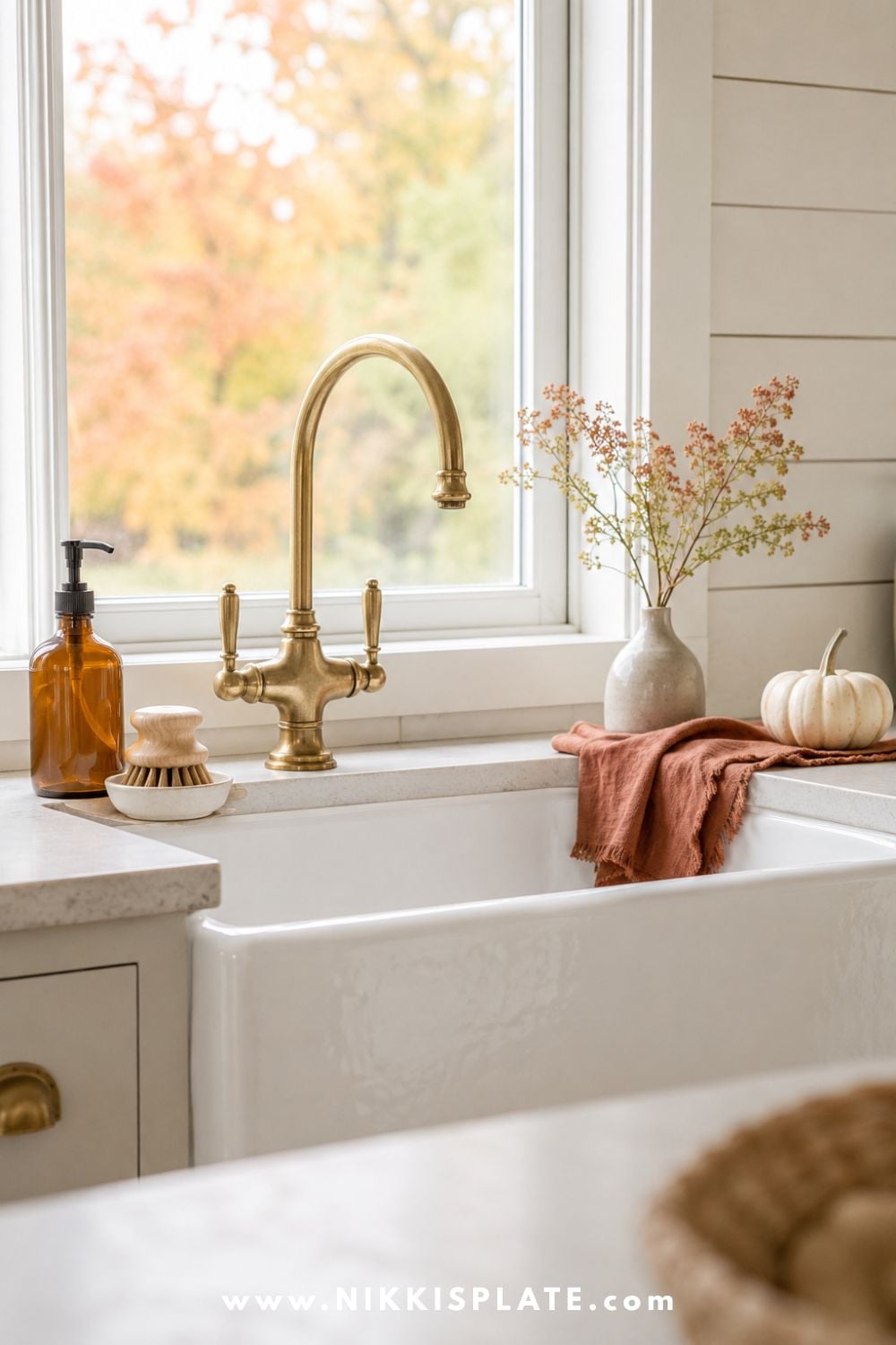 White farmhouse sink decorated for fall with an amber soap dispenser, natural scrub brush, muted rust towel, cream pumpkin and small vase of autumn clippings.