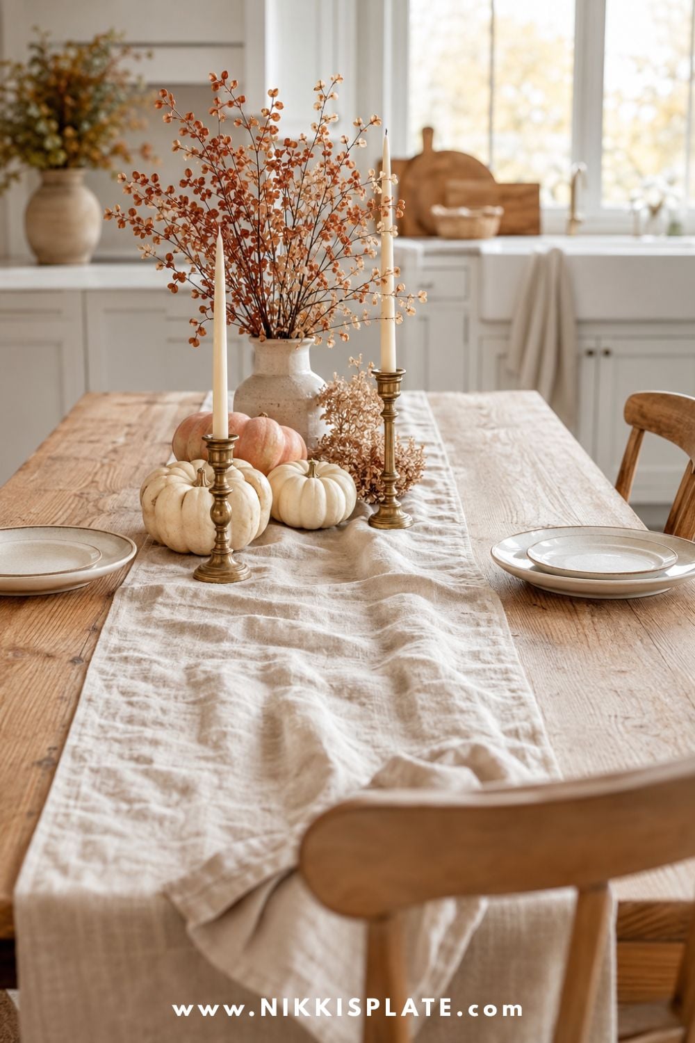 Natural linen runner layered across a warm oak kitchen table with cream pumpkins, faded rust branches, brass candlesticks and simple stoneware place settings.