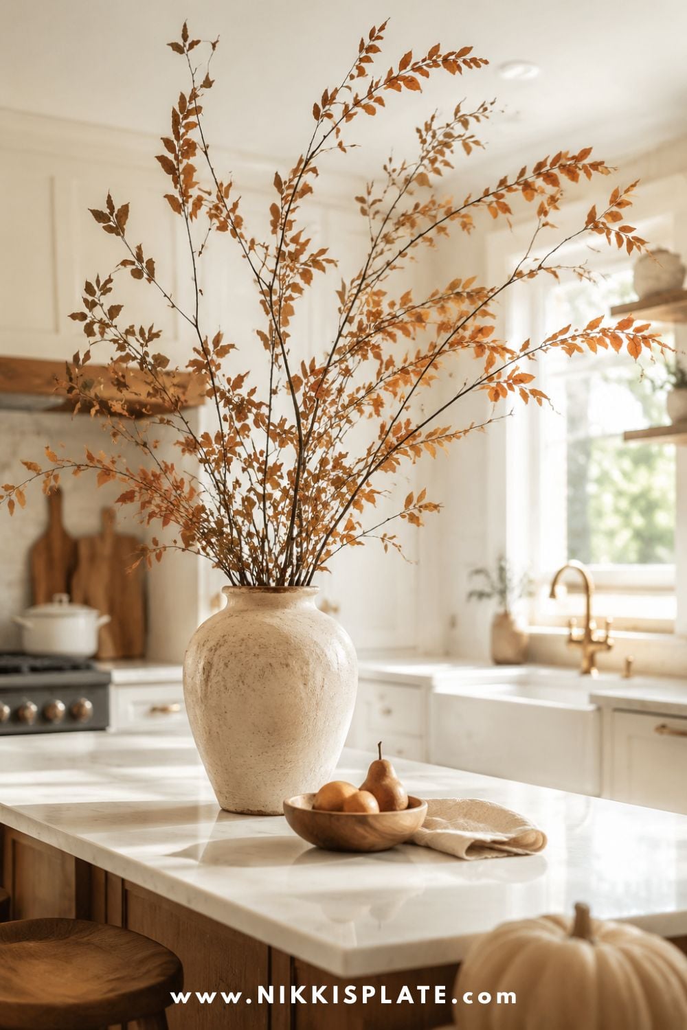 Tall amber and rust fall branches arranged loosely in a cream stoneware vase on a light farmhouse kitchen island beside pears and natural linen.