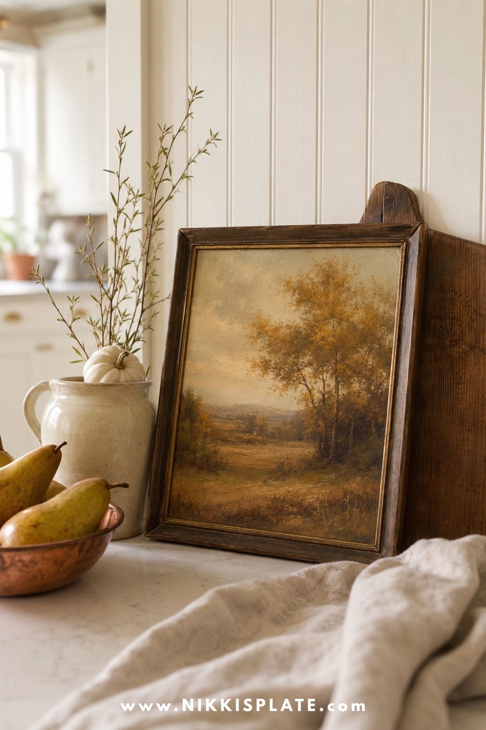 Vintage-style autumn landscape artwork in a dark wood frame styled with a copper bowl of pears, stoneware jug, branches and pale pumpkin on a kitchen counter.