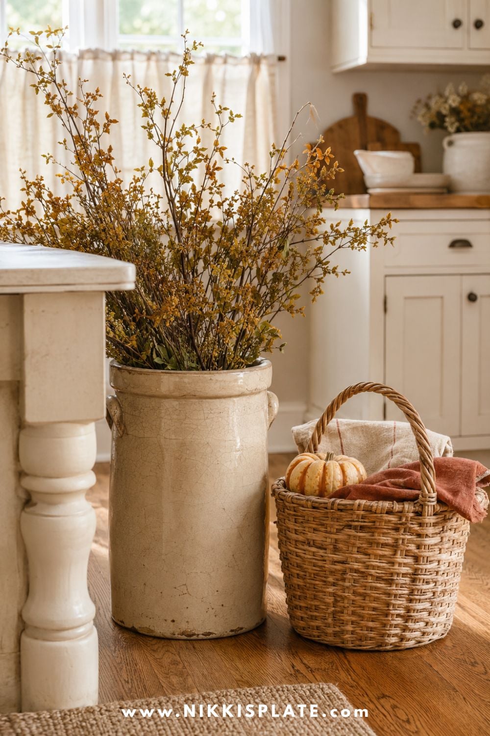 Vintage cream stoneware crock filled with tall autumn branches beside a woven basket holding oatmeal linens, a rust towel and small heirloom pumpkin.