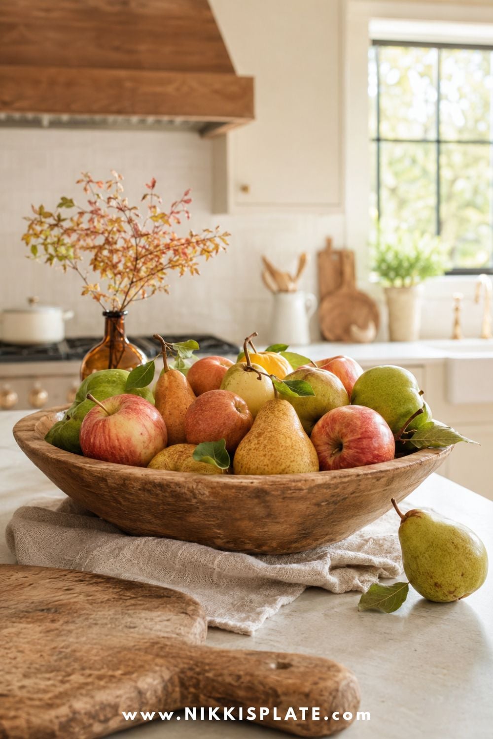 Worn wooden bowl filled with blush-red apples and golden and green pears on a farmhouse kitchen island with an amber bud vase and oatmeal linen.