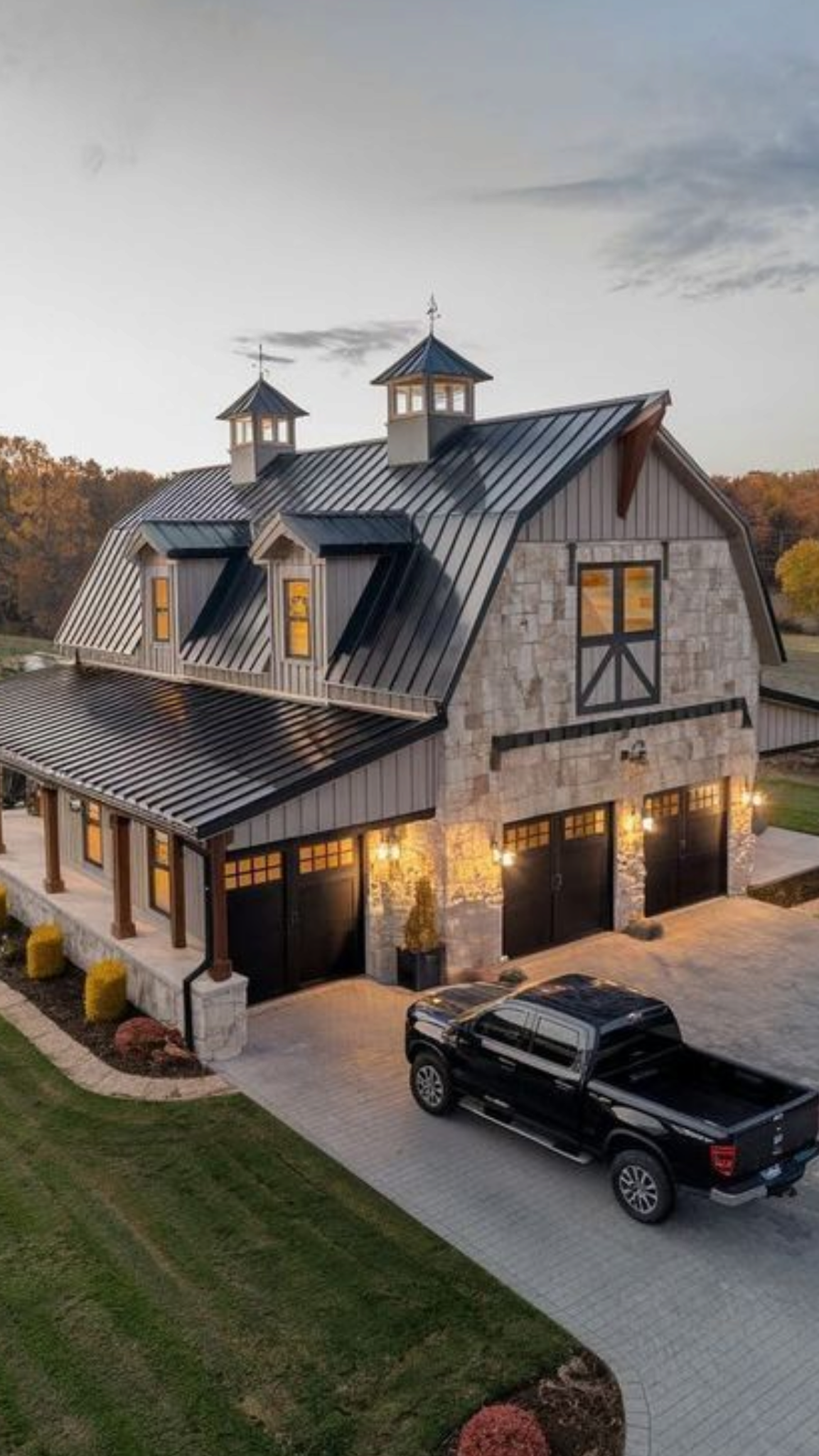 A two-story stone and metal barndominium with black trim and dormer windows, featuring a three-car garage and a covered porch, with warm exterior lighting and a black pickup truck parked in the driveway at sunset.