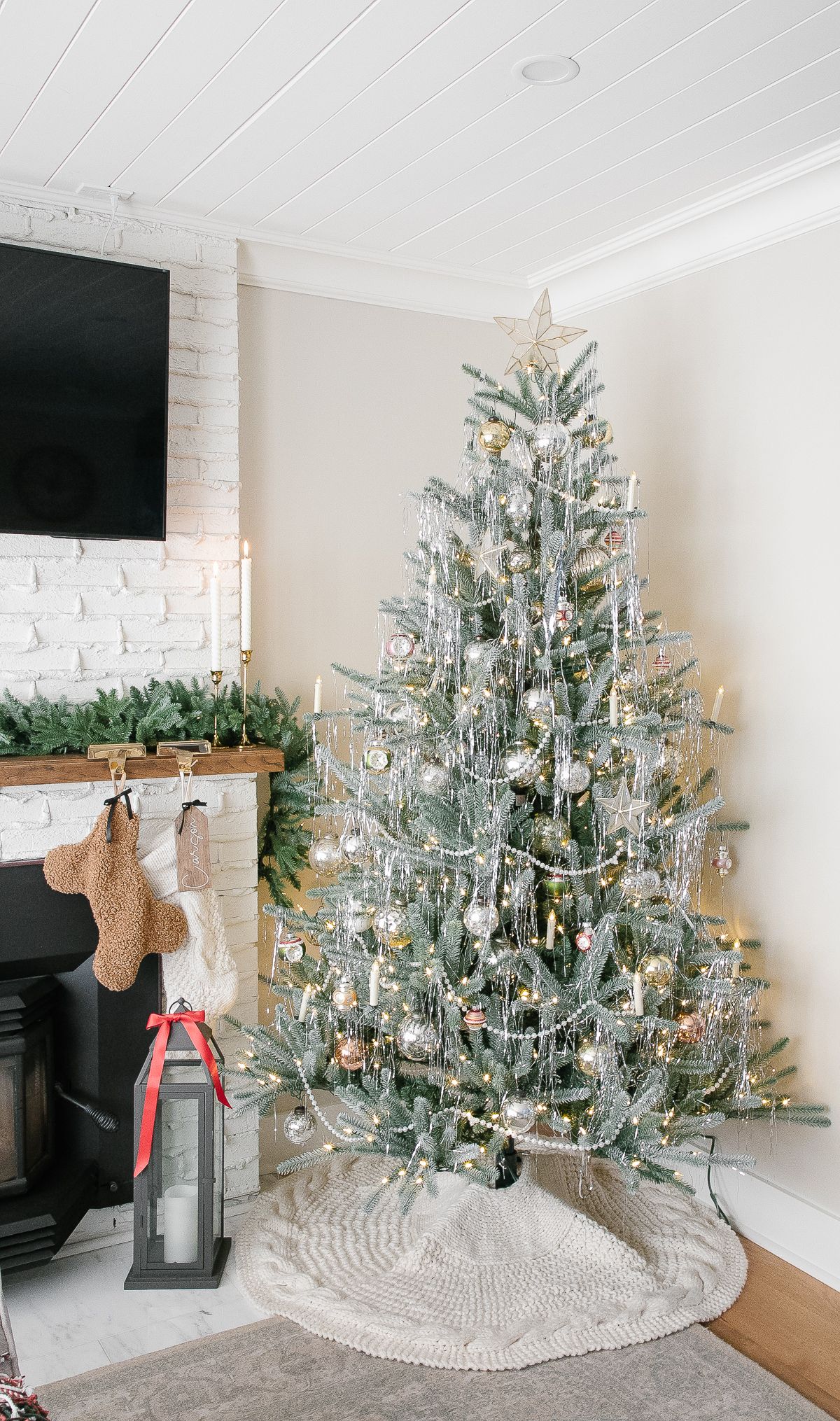 Classic silver tinsel Christmas tree decorated with gold ornaments, white lights, and a cozy knit tree skirt beside a white brick fireplace.