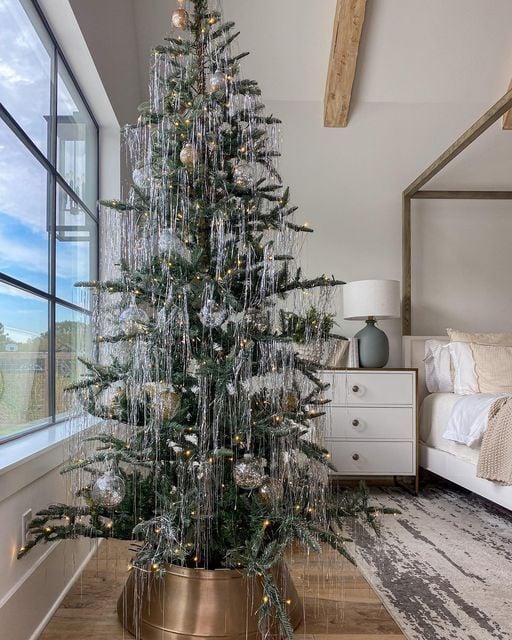 Modern farmhouse tinsel Christmas tree with silver tinsel, gold base, and neutral decor in a bright bedroom with wood beams.