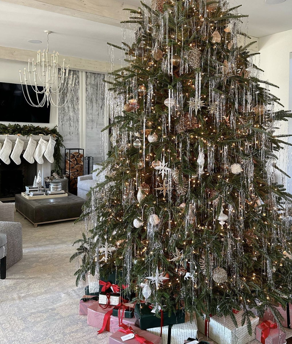 Modern glam tinsel Christmas tree decorated with cascading silver tinsel, metallic ornaments, and wrapped gifts in a cozy living room with stockings on the mantel.