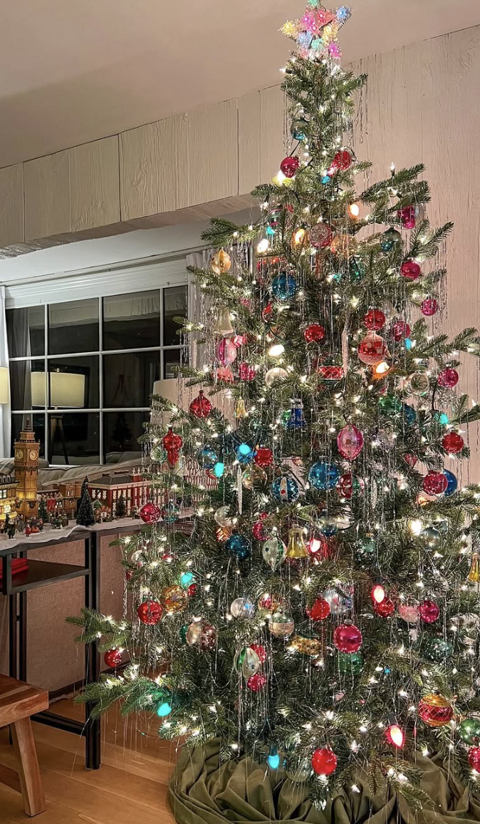 Retro colorful tinsel Christmas tree decorated with silver tinsel, jewel-toned ornaments, and multicolored lights in a cozy living room.