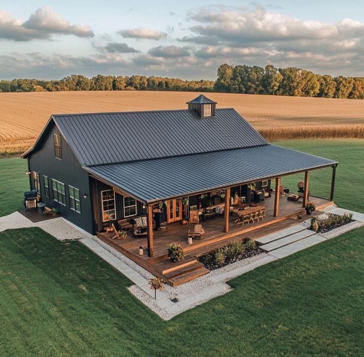 This farmhouse-style black barndominium exudes country charm with its expansive wraparound porch and rich wood details. The dark siding and metal roof contrast beautifully with the surrounding golden fields, creating a picture-perfect rural retreat. The cozy outdoor seating and rustic decor make it an inviting space to relax and enjoy the peaceful countryside.
