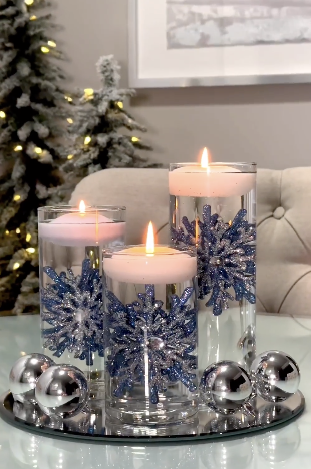 Three glass vases decorated with blue and silver glitter snowflakes topped with floating candles and surrounded by silver ornaments for a winter Christmas display.