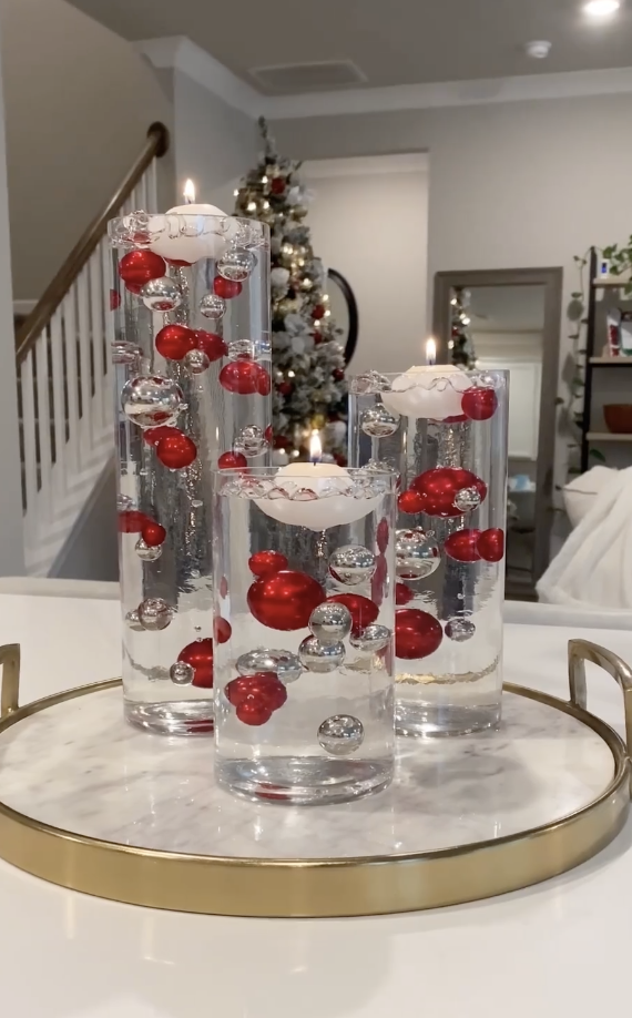 Three tall glass vases filled with water, red and silver ornaments, and floating candles displayed on a marble tray in a holiday-decorated room.