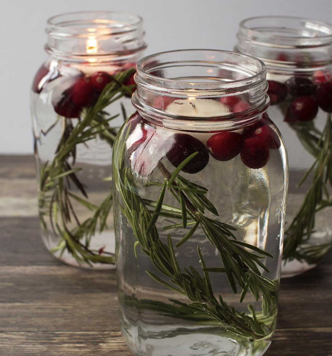 Three mason jars filled with water, rosemary sprigs, and cranberries topped with floating candles for a rustic Christmas centerpiece.