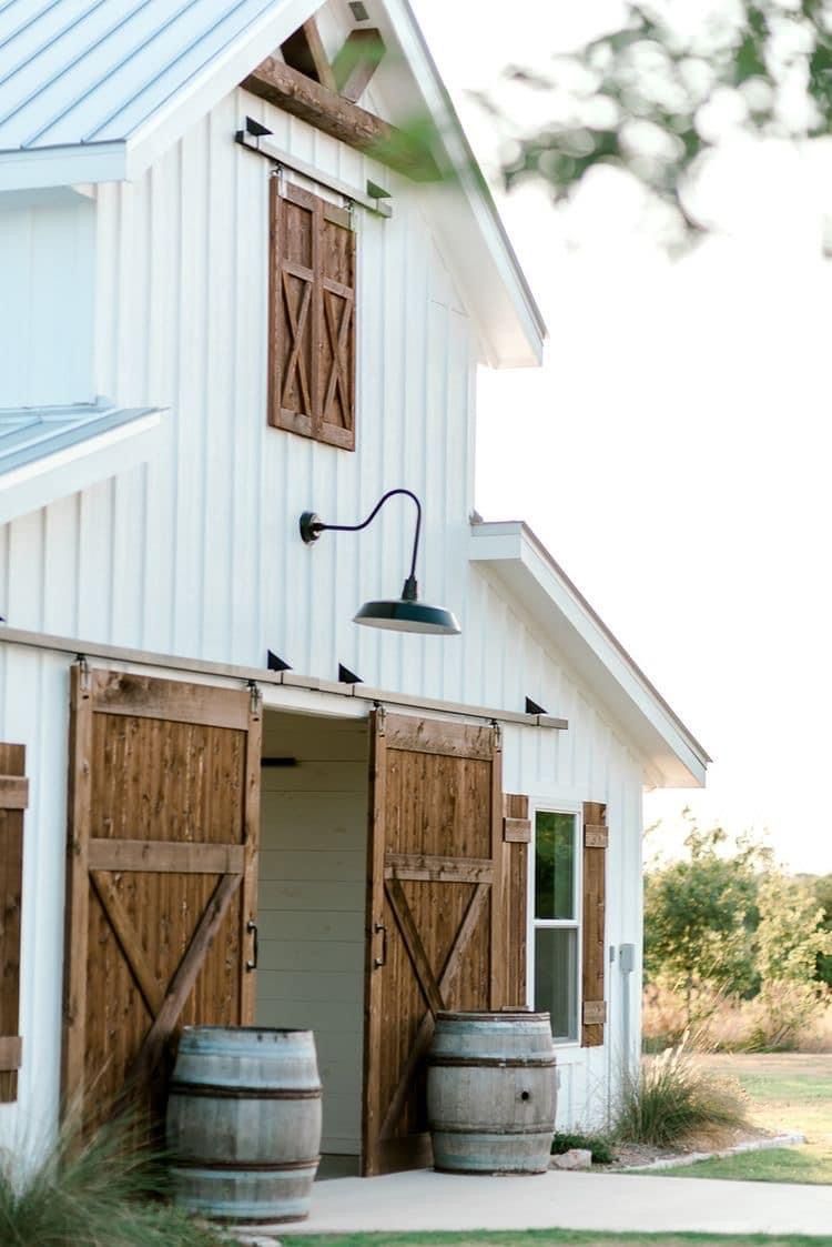 White barndominium with natural wood sliding barn doors, matching shutters, and a black gooseneck light fixture above the entry, accented by decorative wine barrels.