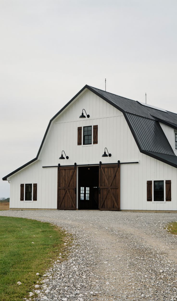 White barndominium with a black metal roof, large sliding wooden barn doors, black window trim, and gooseneck lights along a gravel driveway.