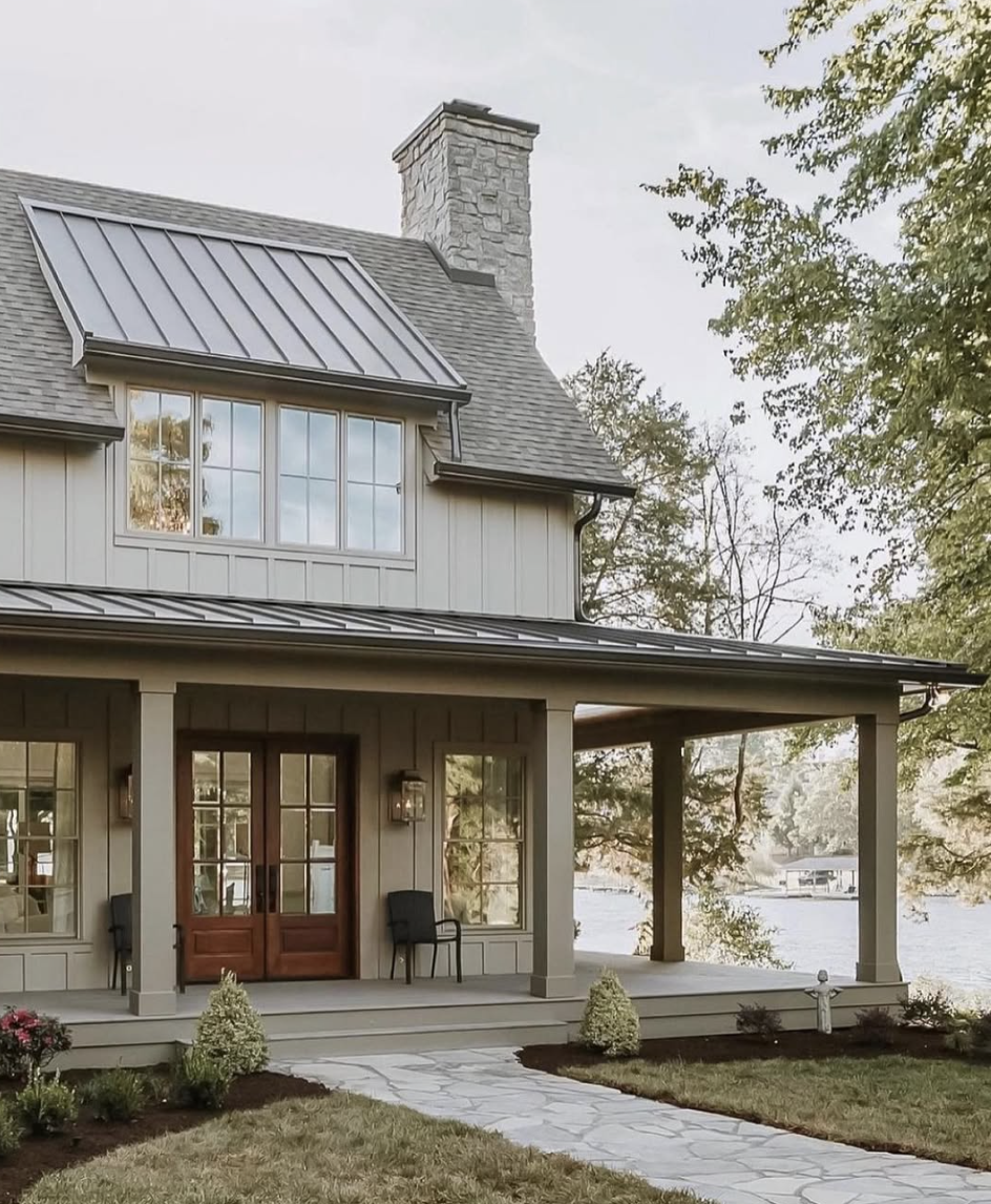 Farmhouse exterior with a standing seam metal roof, warm neutral siding, stone chimney, and covered porch reflecting house exterior trends for 2026.