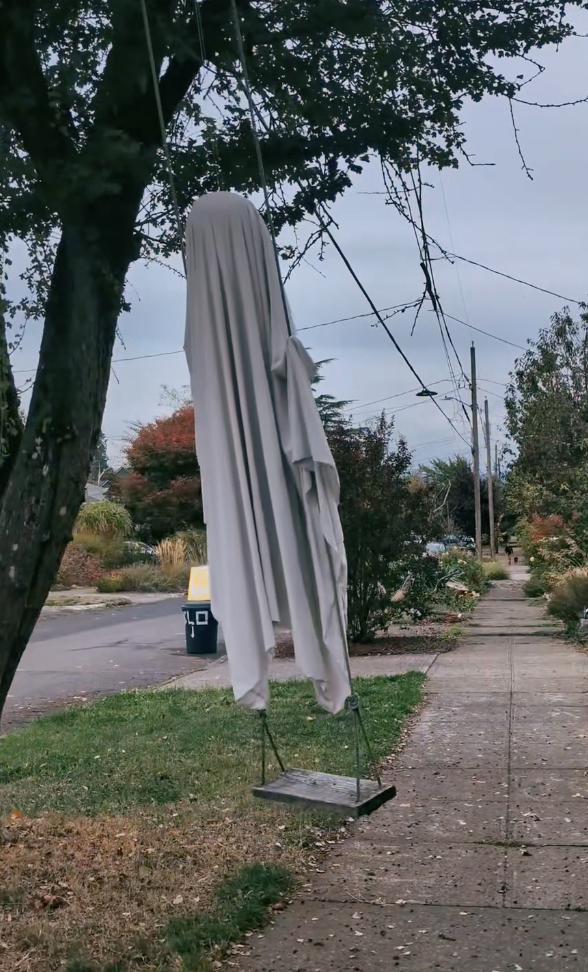 Creepy ghost figure draped in white fabric sitting on a swing hanging from a tree, creating a spooky yard display.