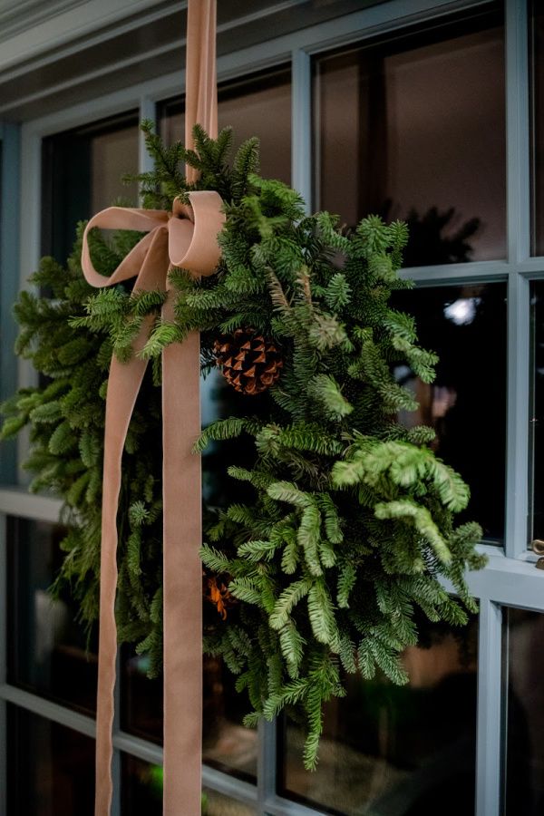 Simple Christmas wreath made of pine greenery and pinecones, hanging by a blush velvet ribbon on a window.