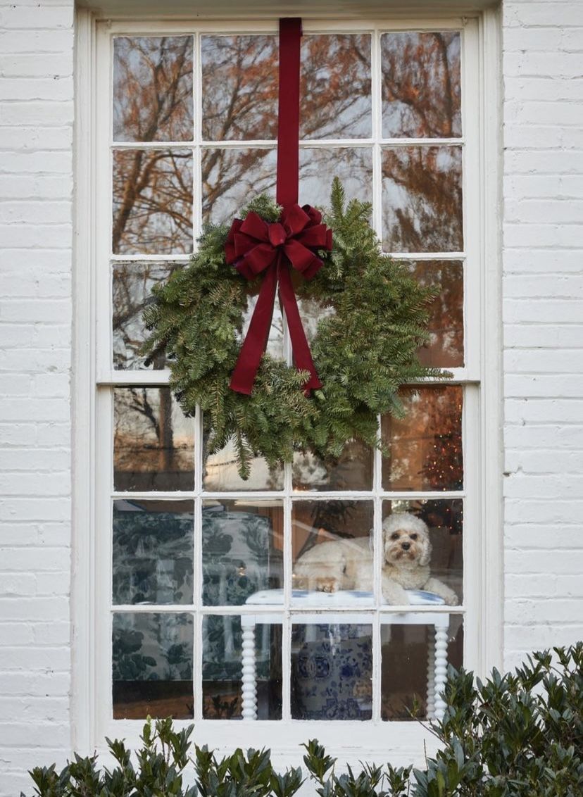 Christmas wreath with burgundy velvet ribbon hanging on a window of a white brick home with a dog sitting inside.