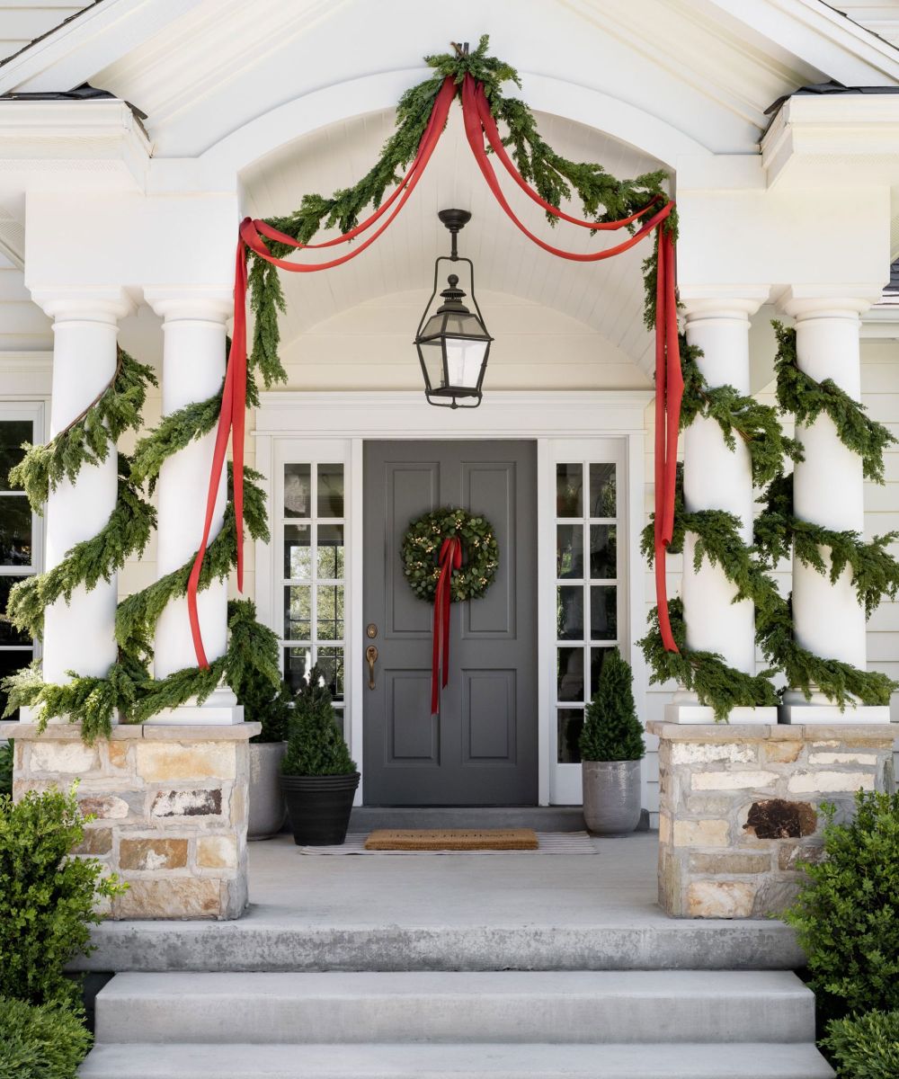 Front porch decorated with red ribbons, lush green garland draped along white columns, and a matching wreath on a dark gray door.