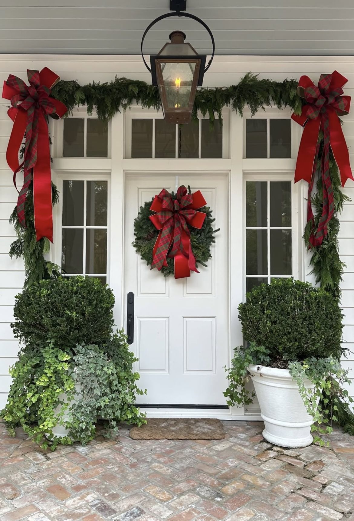 Front porch decorated with red tartan ribbon bows, fresh evergreen garland, and a matching wreath on a white door.