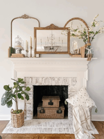This refined summer mantel features a trio of ornate mirrors, brass candlesticks, and soft florals for a look that's both classic and inviting. Soft white blooms spill from a glass jug vase, while stacked vintage books and a cloche add thoughtful, collected charm. Anchored by whitewashed brick and warm wicker textures below, the entire space feels airy, curated, and full of quiet elegance.