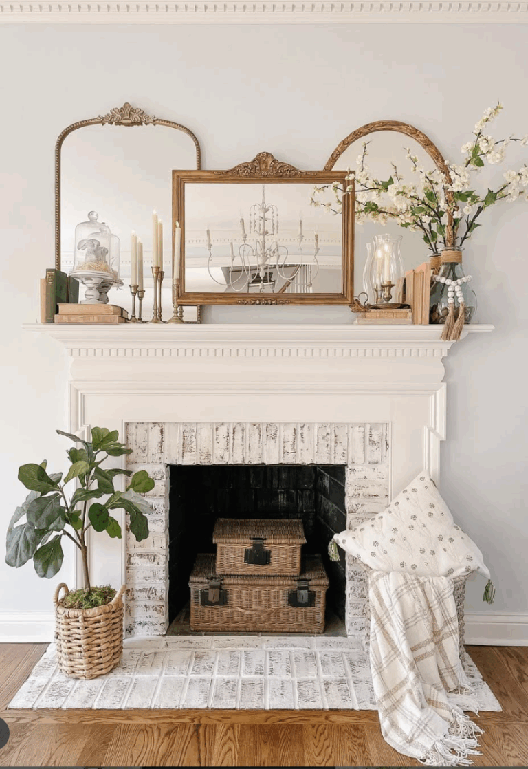 This refined summer mantel features a trio of ornate mirrors, brass candlesticks, and soft florals for a look that's both classic and inviting. Soft white blooms spill from a glass jug vase, while stacked vintage books and a cloche add thoughtful, collected charm. Anchored by whitewashed brick and warm wicker textures below, the entire space feels airy, curated, and full of quiet elegance.