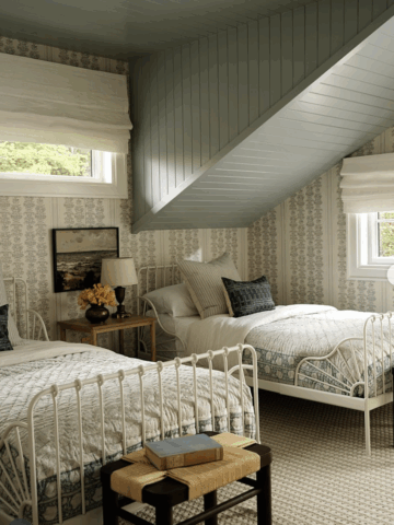 Attic bedroom with two white iron antique twin beds, neutral bedding, vintage wallpaper, a green painted ceiling, and a wooden side table with a lamp and flowers.