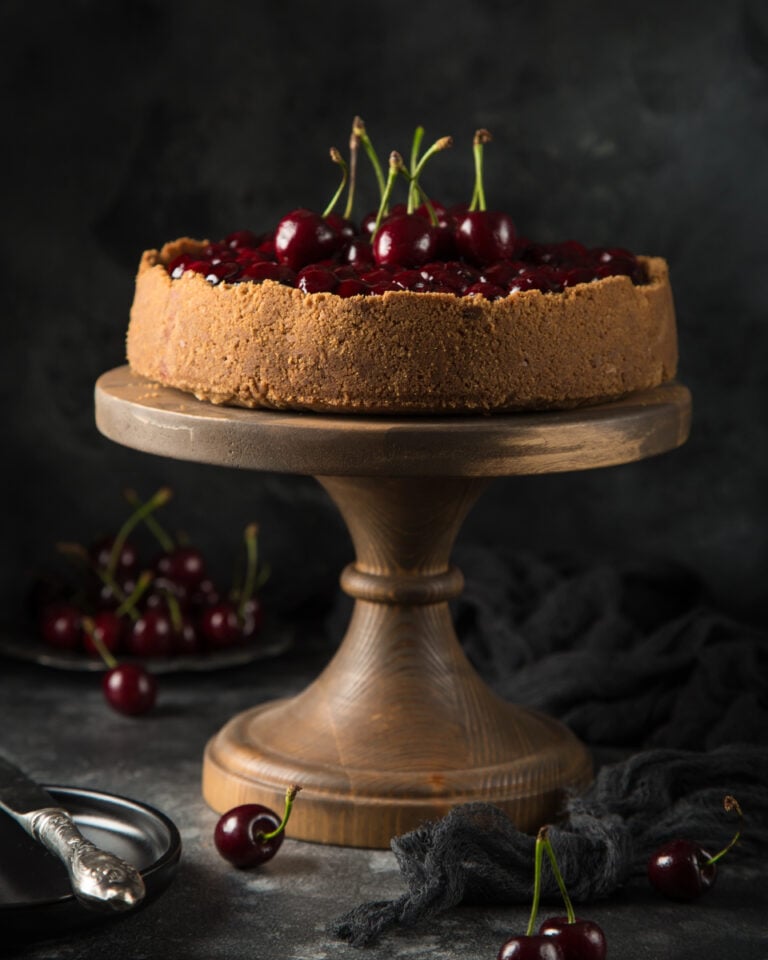 Side view of cherry cheesecake on a wooden cake stand, topped with fresh whole cherries and rustic table decor.