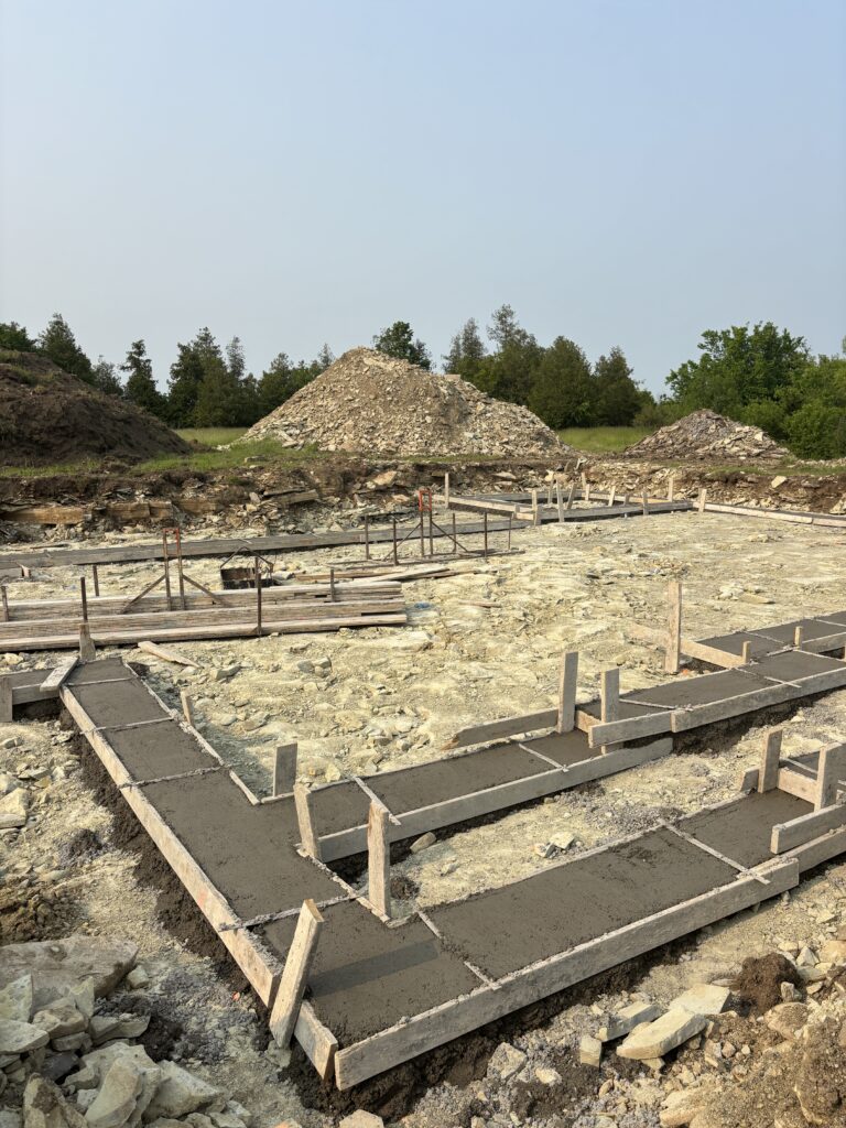 Freshly poured concrete footings in wooden forms, surrounded by rocky soil and construction debris.
