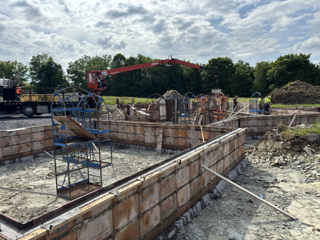Workers pouring concrete into foundation wall forms using a red crane truck during early construction phase.